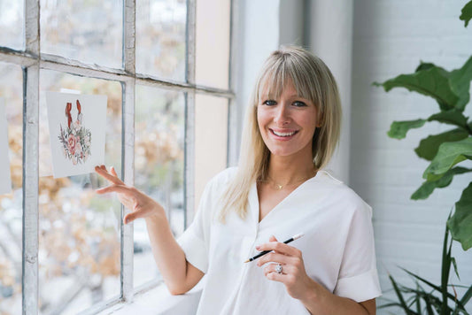 Good Dirt About the artist: Sarah Simon. Sarah Simon standing next to bright sunlit window pointing at her original art.